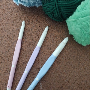 May include: Three crochet hooks with white heads and pastel handles in pink, lavender, and light blue. The handles are marked with sizes 10.0mm, 12.0mm, and 15.0mm. The hooks are positioned near three balls of yarn in blue, dark green, and mint green.