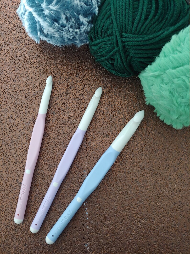 May include: Three crochet hooks with white heads and pastel handles in pink, lavender, and light blue. The handles are marked with sizes 10.0mm, 12.0mm, and 15.0mm. The hooks are positioned near three balls of yarn in blue, dark green, and mint green.