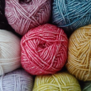 Op de afbeelding: Een verzameling ronde garenbollen in diverse kleuren, waaronder felroze, crème, mosterdgeel, lichtblauw, lavendel en zwart. Elke bol heeft een getwiste vezelstructuur met zichtbare draden die in een dichte, bolvormige structuur zijn gewikkeld.