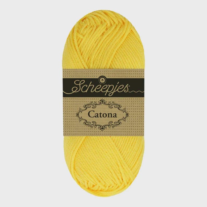 May include: A bright yellow skein of Scheepjes Catona cotton yarn. The yarn is wrapped with a brown label featuring the brand name and the yarn name, Catona, in an ornate font. The yarn is tightly wound and ready for crafting.