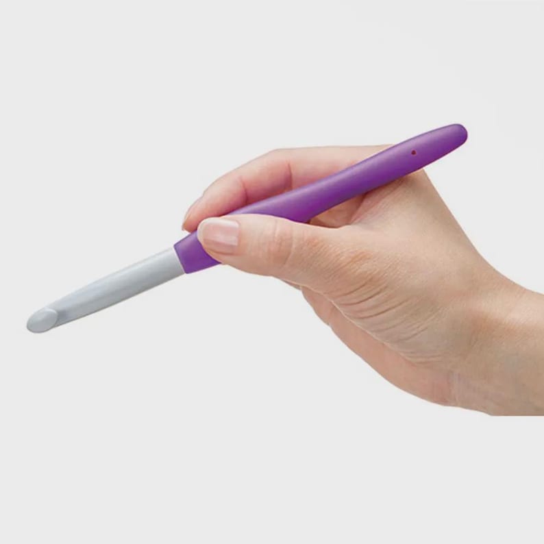 Pode incluir: Uma agulha de croch&ecirc; roxa com uma extremidade de gancho cinza e uma se&ccedil;&atilde;o de cabo branca. A agulha &eacute; segurada em uma m&atilde;o contra um fundo simples. A agulha &eacute; projetada para artesanato com fios.