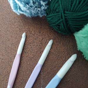 May include: Three crochet hooks with white heads and pastel-colored handles in light pink, lavender, and light blue, positioned alongside three skeins of yarn in dark green, mint green, and a textured blue-grey color.