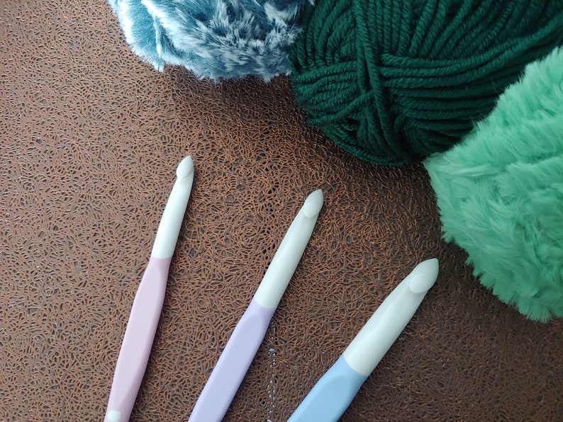 May include: Three crochet hooks with white heads and pastel-colored handles in light pink, lavender, and light blue, positioned alongside three skeins of yarn in dark green, mint green, and a textured blue-grey color.