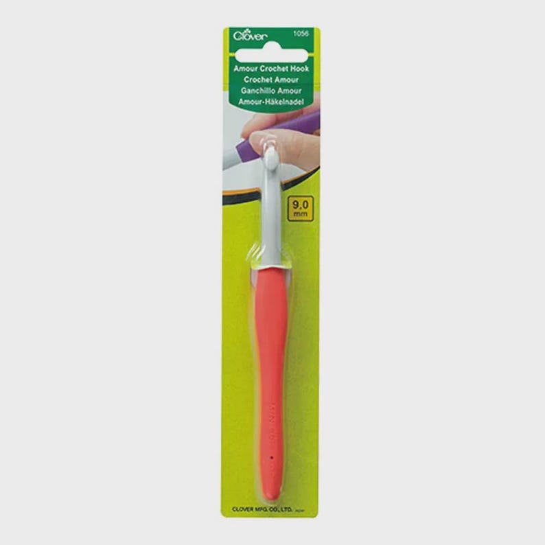 Pode incluir: Uma agulha de croch&ecirc; com cabo vermelho e ponta branca, embalada em um cart&atilde;o verde e amarelo. O cart&atilde;o exibe o texto "Amour Crochet Hook" e o tamanho "9,0 mm". O logotipo da marca Clover &eacute; vis&iacute;vel no topo.