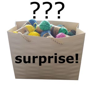 May include: A white basket overflowing with colourful yarn skeins in shades of blue, green, yellow, and pink. The basket has rope handles and the word "surprise!" is printed on the front, with question marks above.