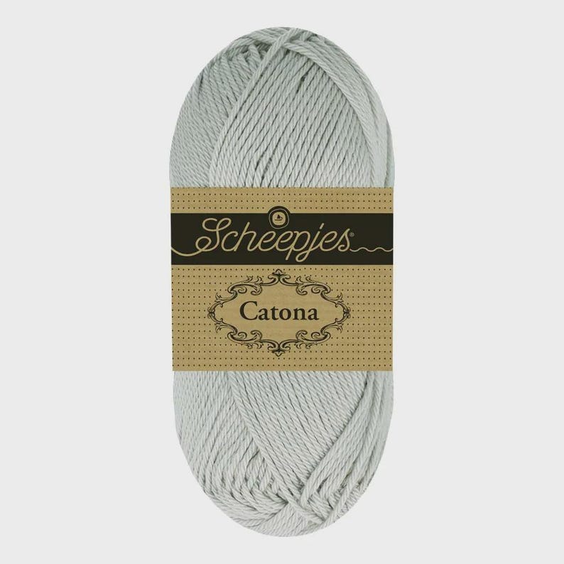 May include: A skein of light gray yarn with a brown label. The label features the brand name "Scheepjes" in cursive and the word "Catona" in a decorative frame. The yarn is tightly wound.