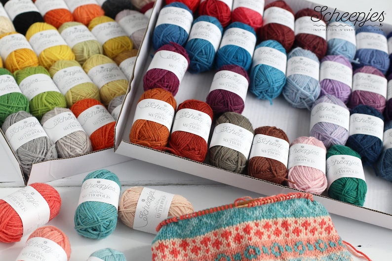 May include: A variety of colorful yarn skeins in different shades of red, orange, yellow, green, blue, purple, brown, and pink. The yarn is arranged in two boxes and on a white surface. The yarn is labeled "Scheepjes" and "Wool".