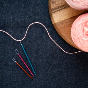 May include: Three knitting needles, one blue, one red, and one orange, are laid out on a dark gray surface. A pink yarn is looped around the needles and a ball of pink yarn is on a wooden surface.