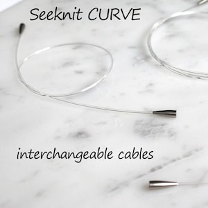 May include: Seeknit CURVE interchangeable cables for knitting. The cables are silver and have a smooth finish. The cables are shown on a white marble surface.