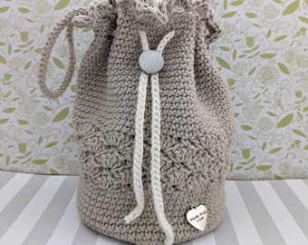 Puede incluir: Un bolso beige de ganchillo con cordón y un patrón decorativo. El bolso tiene un cordón blanco y un cierre de botón plateado. Un pequeño dije en forma de corazón con la inscripción "Made with Love" cuelga del bolso.