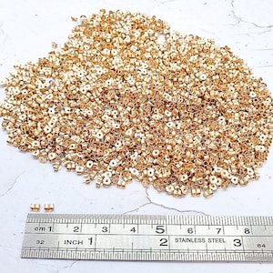 May include: A large pile of small, gold-colored earring backs. A stainless steel ruler with inch and centimeter markings is in the foreground. The earring backs are scattered across a white surface.