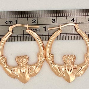 May include: A pair of rose gold hoop earrings with a Celtic knot design featuring a crown and heart. The earrings are 2.5 cm in diameter.