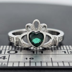 Puede incluir: Un anillo Claddagh plateado con una piedra preciosa verde esmeralda en forma de corazón. El anillo presenta una corona sobre el corazón y manos que lo abrazan. El anillo está sobre una superficie negra junto a una regla.