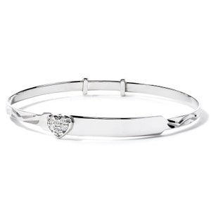 May include: A silver-toned baby bangle bracelet. The bracelet features a heart-shaped charm set with small, clear gemstones. The band has a smooth, adjustable design with decorative etched details near the charm.