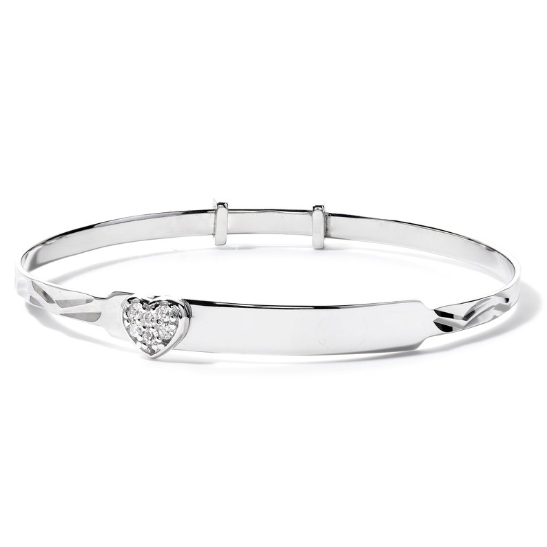 May include: A silver-toned baby bangle bracelet. The bracelet features a heart-shaped charm set with small, clear gemstones. The band has a smooth, adjustable design with decorative etched details near the charm.