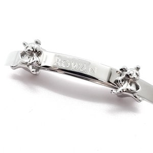 May include: Silver-toned baby bracelet with the name "ROWAN" engraved on a curved bar. Two small teddy bear figures are attached to the bracelet, one on each side of the name. The bracelet is designed for a baby.