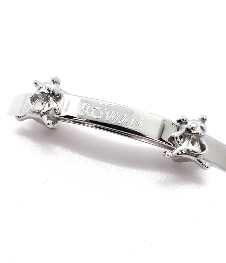 May include: Silver-toned baby bracelet with the name "ROWAN" engraved on a curved bar. Two small teddy bear figures are attached to the bracelet, one on each side of the name. The bracelet is designed for a baby.