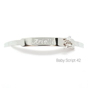 May include: A silver baby bracelet with the name "Arielle" engraved on a rectangular plate. A small teddy bear charm is attached to the bracelet. The text "Baby Script 42" is below the bracelet.