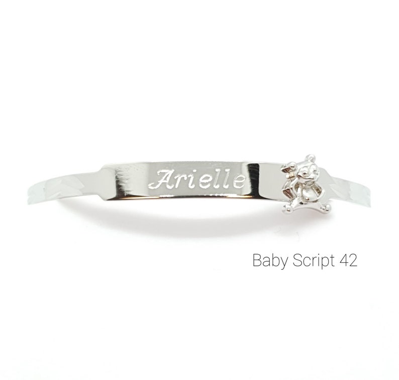 May include: A silver baby bracelet with the name "Arielle" engraved on a rectangular plate. A small teddy bear charm is attached to the bracelet. The text "Baby Script 42" is below the bracelet.