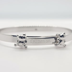 May include: A silver-colored bangle bracelet with the name "Sebastian" engraved on it. The bracelet features two small bear-shaped charms on either side of the name. The bracelet is made of a shiny, reflective metal.