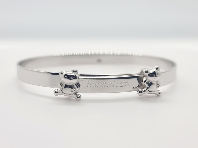 May include: A silver-colored bangle bracelet with the name "Sebastian" engraved on it. The bracelet features two small bear-shaped charms on either side of the name. The bracelet is made of a shiny, reflective metal.