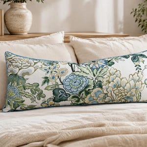 May include: A rectangular lumbar cushion featuring a white base with a detailed botanical print in shades of blue, green, and soft yellow. The cushion is finished with a thin blue piped edge, showcasing large floral motifs and intricate foliage patterns.