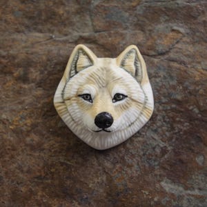 May include: A sculpted wolf head brooch featuring a cream and tan colour palette with detailed fur texture, pointed ears, dark almond-shaped eyes, and a black nose. The wolf face is rendered with realistic shading and defined facial features.