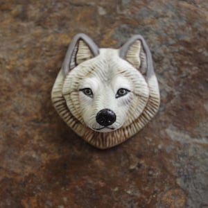 May include: A sculpted wolf head brooch featuring detailed fur textures in shades of white, cream, and light brown. The wolf has pointed ears with grey accents, dark almond-shaped eyes, and a prominent black nose, creating a realistic animal portrait.