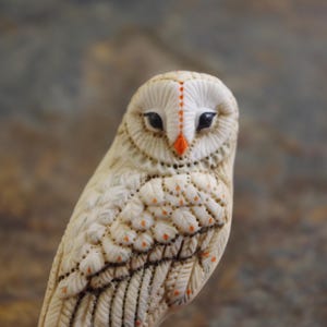 以下が含まれることがあります： 複雑な羽の彫刻が施されたオフホワイトのフクロウの置物。黒い目と鮮やかなオレンジ色のくちばしが特徴です。顔の中央にはオレンジ色のドットの縦線があり、翼には小さなオレンジ色のアクセントが施されています。