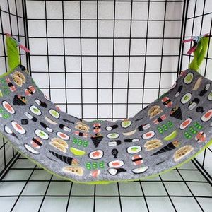 May include: A gray fabric hammock for small pets featuring a repeating pattern of colorful sushi rolls, nigiri, and edamame. The hammock has bright lime green fabric corners and is suspended by pink plastic clips within a black wire cage frame.