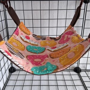 May include: A pink pet hammock with a donut pattern. The hammock is suspended by brown straps and features various colorful donuts with sprinkles and frosting. The background is a black wire cage.