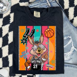 May include: A black t-shirt featuring a cartoon coyote wearing star-shaped glasses and a basketball jersey with the number 2. The graphic includes a basketball, the text GO SPURS GO, and the San Antonio Spurs logo against vertical stripes.