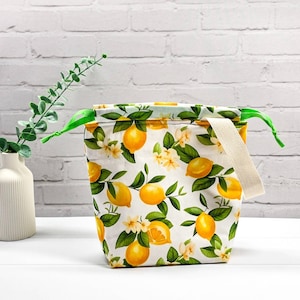 Könnte beinhalten: Eine Lunchtasche aus Stoff mit weißem Hintergrund und einem wiederkehrenden Muster aus gelben Zitronen, grünen Blättern und kleinen weißen Blüten. Die Tasche verfügt über einen beigen Webgriff und leuchtend grüne Kordelzüge oben.