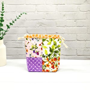 Könnte beinhalten: Ein Patchwork-Stoffbeutel mit Kordelzug und floralen und geometrischen Mustern. Die Tasche hat eine Kombination aus rosa, lila, weißen und orangefarbenen Stoffen. Der obere Rand ist mit orangefarbenem Blumenstoff gefüttert, und die Tasche ist mit einem cremefarbenen Kordelzug gebunden.