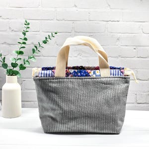 Könnte beinhalten: Eine graue Cord-Tragetasche mit zwei cremefarbenen Stoffgriffen. Das Innere verfügt über ein gestepptes Futter mit einem bunten Patchwork-Muster aus blauen, violetten und weißen Blumen- und Vichy-Karos, gesichert mit dünnen cremefarbenen Kordelzügen.