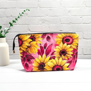 Könnte beinhalten: Eine rechteckige Kosmetiktasche mit einem lebendigen Blumenmuster. Die Tasche zeigt große gelbe Sonnenblumen mit dunkelroten Zentren und burgunderfarbenen Blättern auf einem rosa Hintergrund. Sie hat einen schwarzen Reißverschluss.