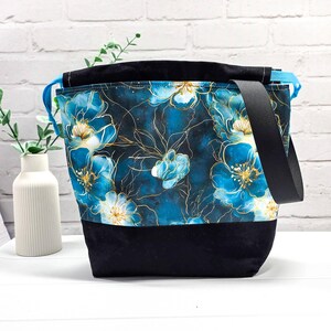 Könnte beinhalten: Ein Beutel mit einem dunkelblauen und türkisfarbenen Blumenmuster mit goldenen Linienakzenten im oberen Bereich und einem einfarbigen schwarzen Boden. Die Tasche verfügt über einen schwarzen Schultergurt und hellblaue Kordelzüge.