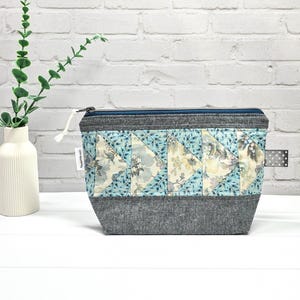 Könnte beinhalten: Eine gesteppte Kosmetiktasche mit blauem und floralem Muster. Die Tasche hat einen grauen Boden und einen Reißverschluss. Auf der Seite ist ein weißes Etikett angebracht. Ein Zweig Grün in einer weißen Vase befindet sich im Hintergrund.