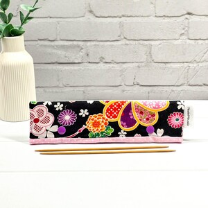 Könnte beinhalten: Schwarzes Stoff-Stricknadel-Etui mit einem Blumenmuster in Rosa, Orange, Lila und Weiß. Das Etui hat einen rosa Rand und wird mit zwei lila Druckknöpfen verschlossen. Zwei Stricknadeln aus Holz sind sichtbar.