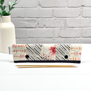 May include: A fabric knitting needle case with a patchwork design in cream, black, pink, and orange. The case is closed with two black press studs and contains two wooden knitting needles. The case is on a white surface.