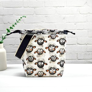 Könnte beinhalten: Eine weiße Stofftasche mit einem sich wiederholenden Muster von Cartoon-Schafen. Die Schafe haben schwarze Gesichter, flauschige weiße Wolle und große, ausdrucksstarke Augen. Die Tasche hat einen schwarzen Riemen und ein schwarzes Innenfutter.