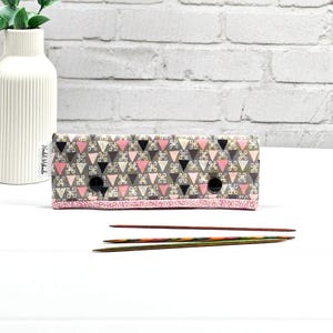 May include: A rectangular knitting needle case with a geometric pattern of pink, black, and grey triangles. The case has two black holes and a pink border. Three knitting needles are in front of the case.