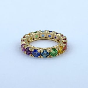 Può includere: Un anello d'oro con una fascia di pietre preziose colorate. L'anello presenta un arcobaleno di colori, tra cui rosso, arancione, giallo, verde, blu e viola. Le pietre sono uniformemente distanziate.