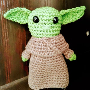 K&ouml;nnte beinhalten: Handgemachte H&auml;kelpuppe einer Figur mit gro&szlig;en gr&uuml;nen Ohren, schwarzen Augen und einem hellgr&uuml;nen Gesicht. Die Puppe tr&auml;gt eine beige Robe und hat kleine gr&uuml;ne H&auml;nde. Die Puppe steht auf einer dunklen Oberfl&auml;che.