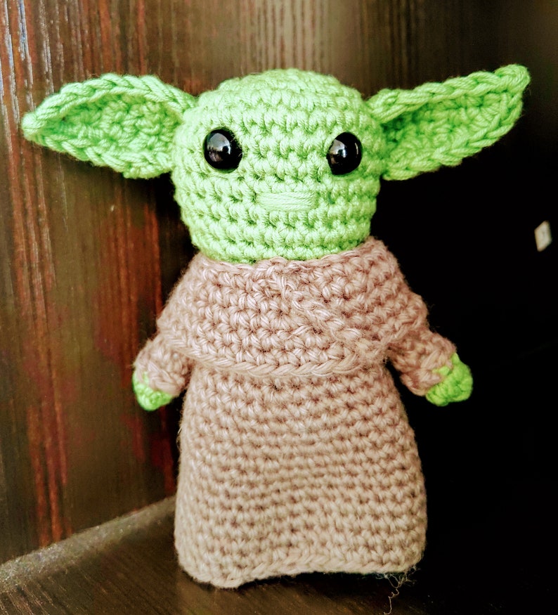 K&ouml;nnte beinhalten: Handgemachte H&auml;kelpuppe einer Figur mit gro&szlig;en gr&uuml;nen Ohren, schwarzen Augen und einem hellgr&uuml;nen Gesicht. Die Puppe tr&auml;gt eine beige Robe und hat kleine gr&uuml;ne H&auml;nde. Die Puppe steht auf einer dunklen Oberfl&auml;che.