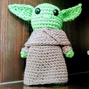 K&ouml;nnte beinhalten: Handgemachte H&auml;kelpuppe einer Figur mit gro&szlig;en gr&uuml;nen Ohren und schwarzen Augen. Die Puppe tr&auml;gt eine beige Robe und hat gr&uuml;ne H&auml;nde und F&uuml;&szlig;e. Die Puppe steht aufrecht und ist aus Garn gefertigt.