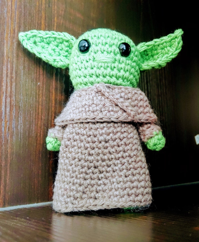 K&ouml;nnte beinhalten: Handgemachte H&auml;kelpuppe einer Figur mit gro&szlig;en gr&uuml;nen Ohren und schwarzen Augen. Die Puppe tr&auml;gt eine beige Robe und hat gr&uuml;ne H&auml;nde und F&uuml;&szlig;e. Die Puppe steht aufrecht und ist aus Garn gefertigt.