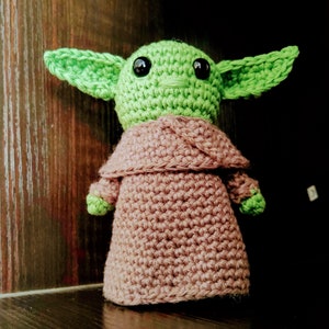 K&ouml;nnte beinhalten: Handgemachte H&auml;kelpuppe einer Figur mit gro&szlig;en gr&uuml;nen Ohren, einem gr&uuml;nen Kopf und schwarzen Augen. Die Puppe tr&auml;gt eine braune Robe. Die Puppe ist etwa 15 cm gro&szlig;.