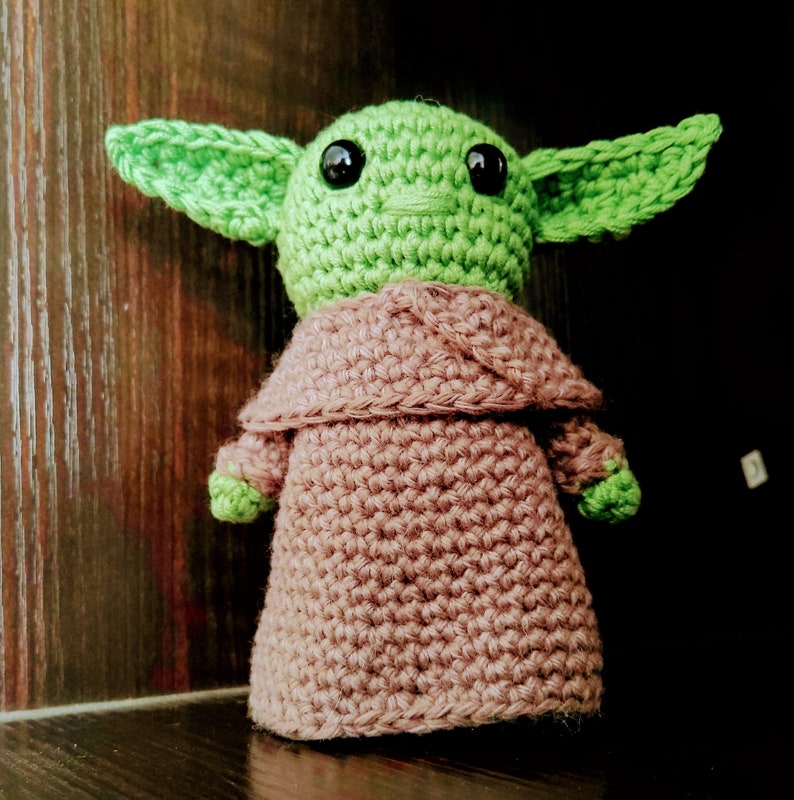 K&ouml;nnte beinhalten: Handgemachte H&auml;kelpuppe einer Figur mit gro&szlig;en gr&uuml;nen Ohren, einem gr&uuml;nen Kopf und schwarzen Augen. Die Puppe tr&auml;gt eine braune Robe. Die Puppe ist etwa 15 cm gro&szlig;.