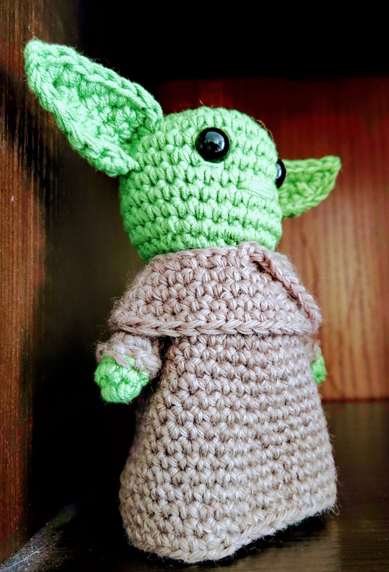 K&ouml;nnte beinhalten: Handgefertigte H&auml;kelpuppe einer Figur mit gro&szlig;en Ohren und schwarzen Augen. Die Puppe ist haupts&auml;chlich gr&uuml;n mit einem beigen Umhang. Die Puppe ist eine beliebte Figur aus einem Science-Fiction-Franchise.